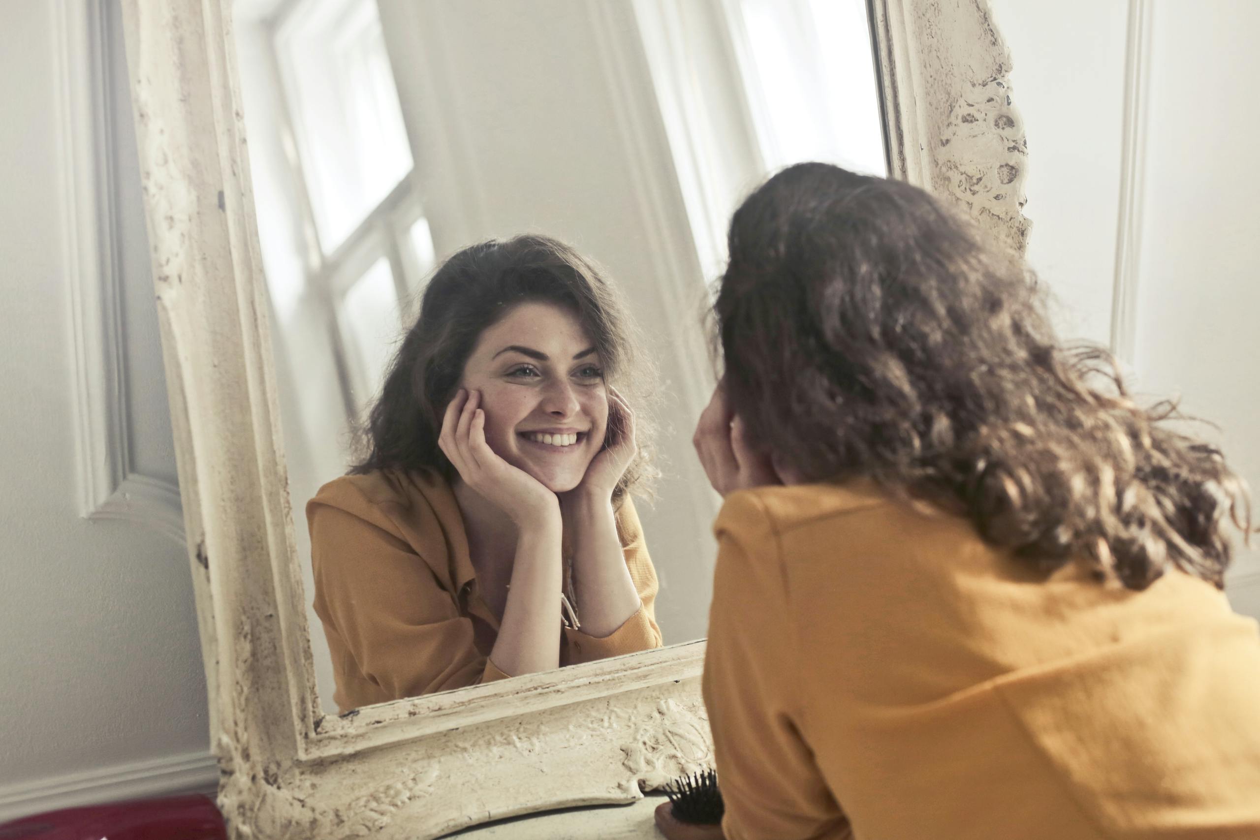 The Body Project 1 A cheerful woman smiles at her reflection in a vintage-style mirror, exuding positivity and warmth.