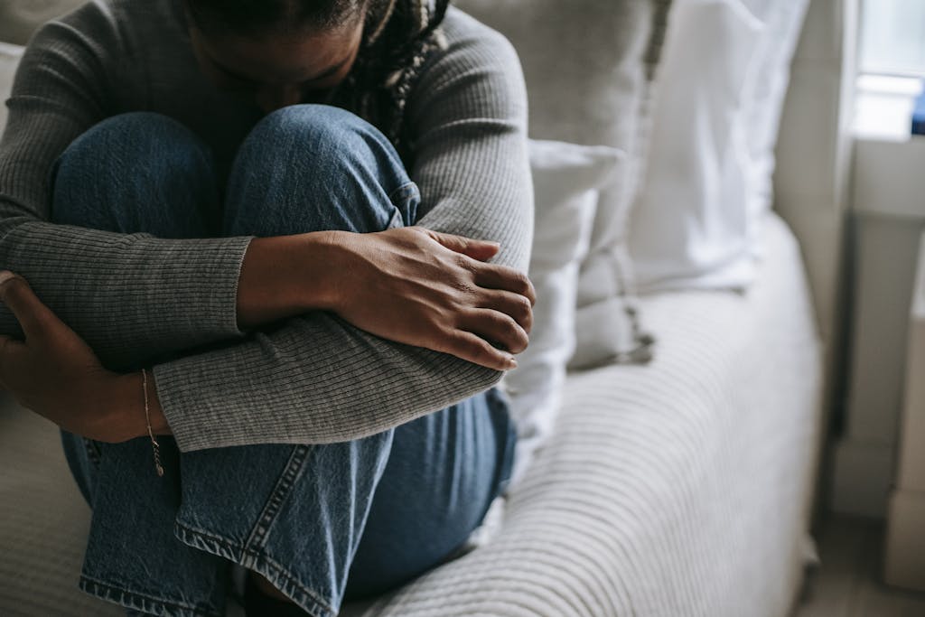 A woman sits on a bed, hugging her knees in a contemplative pose, indoors.